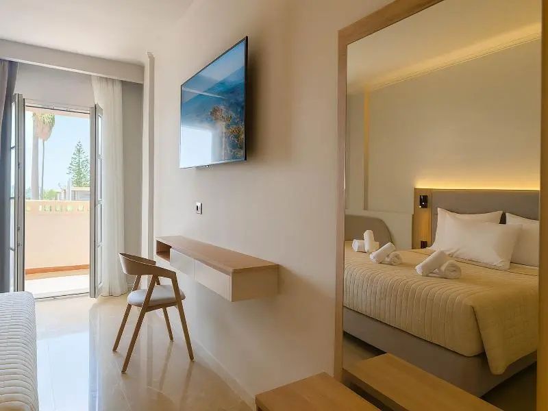Helles Hotelzimmer mit Doppelbett im Spiegel, Wandfernseher, Holzschreibtisch und Zugang zum Balkon.