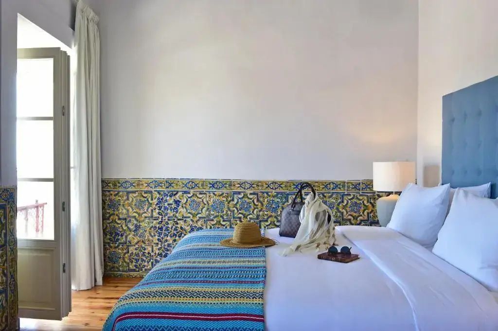 Hotelbett mit weißer Bettwäsche, bunten Kissen, Strohhut und einer Wand aus gelb-blauen Fliesen.