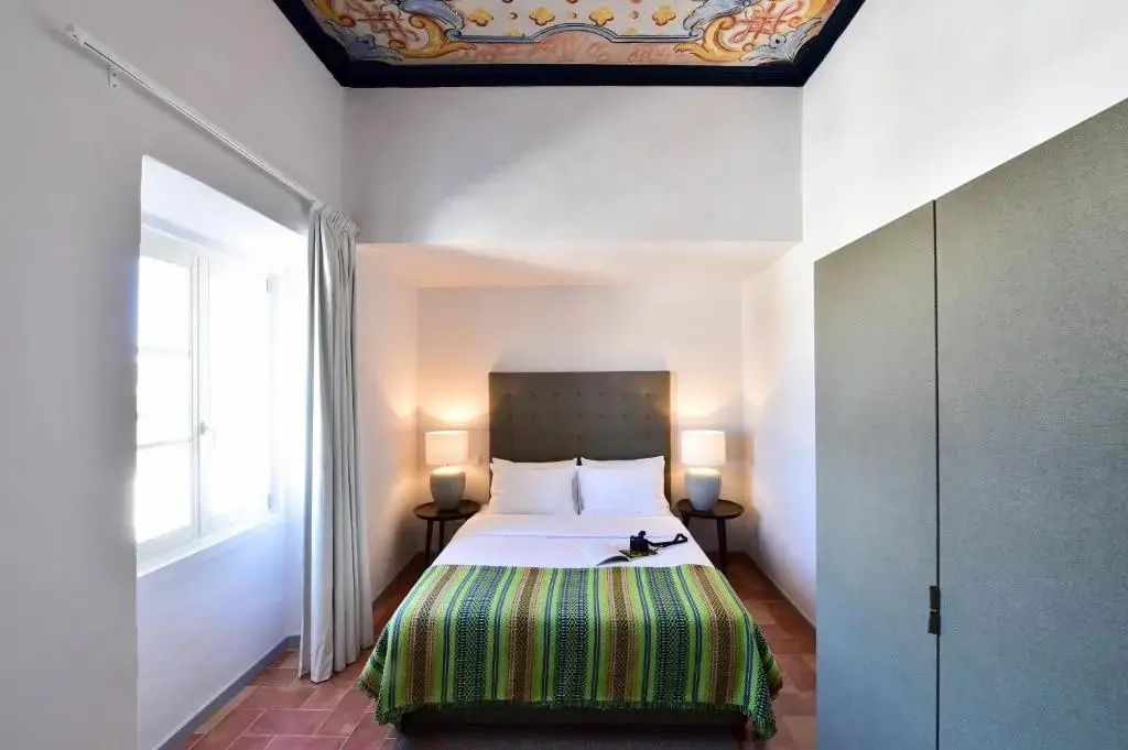 Hell eingerichtetes Hotelzimmer mit bemaltem Deckenfresko und Doppelbett.