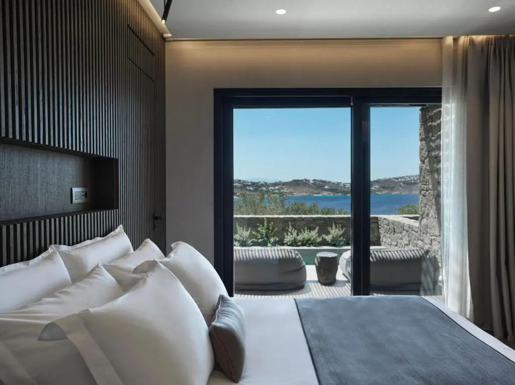 Doppelbett mit weißen Kissen und Blick durch die Balkontür auf das Meer und die Terrasse.
