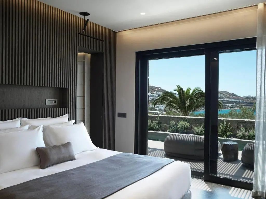 Doppelbett in einem modern gestalteten Hotelzimmer mit Blick durch die Glasfront auf eine Terrasse.
