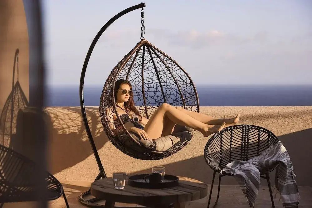 Eine Frau sitzt in einem braunen Hängesessel auf einer Terrasse mit Blick auf das Meer.