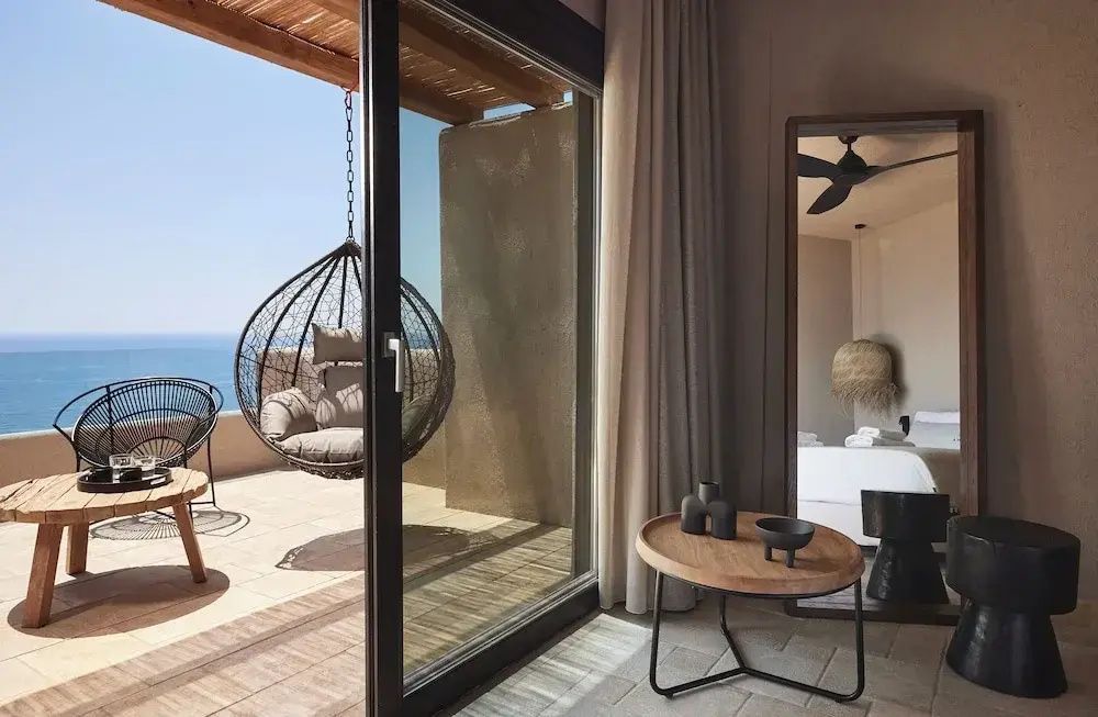Blick aus dem Zimmer durch eine Glastür auf die Terrasse mit Hängesessel und das Meer.