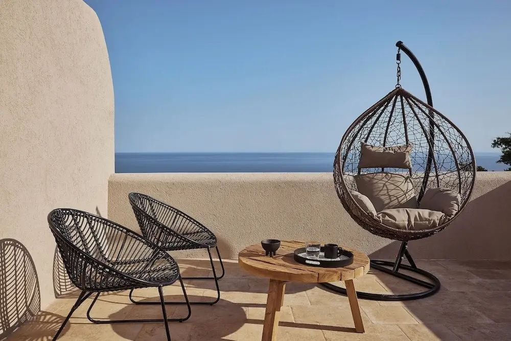 Sonnige Terrasse mit zwei schwarzen Stühlen, Holztisch und Hängesessel vor dem Meer.