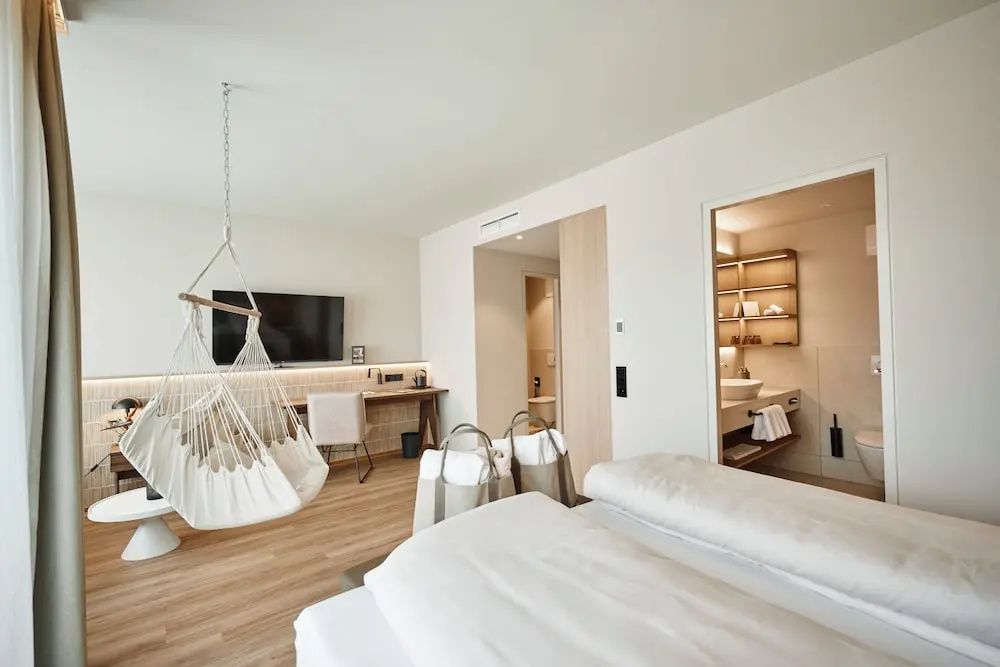 Helles Hotelzimmer mit Doppelbett, Hängesessel, Schreibtisch und Blick ins angrenzende Badezimmer.