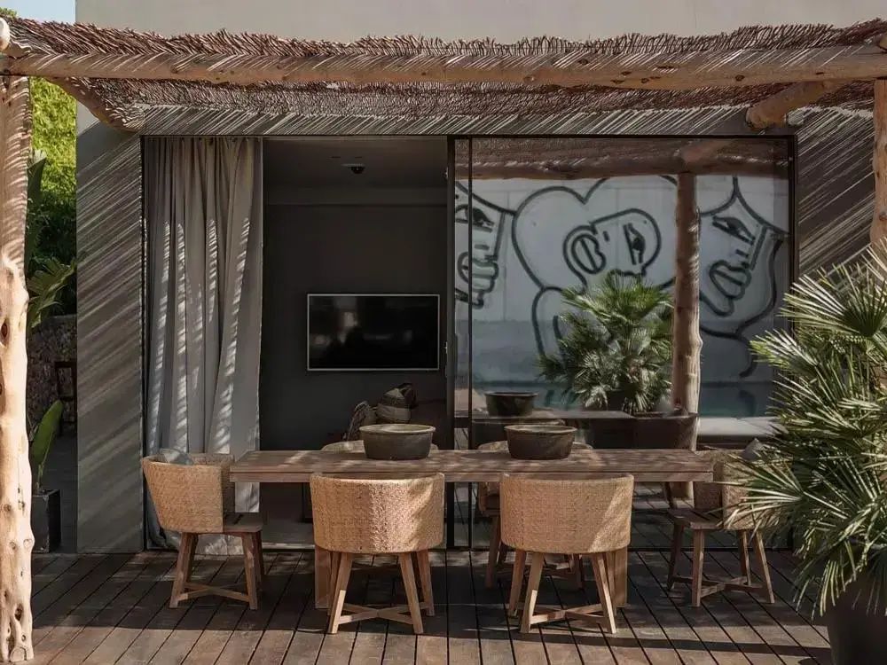 Holztisch mit geflochtenen Stühlen auf einer Holzterrasse unter einer Pergola aus Reet.