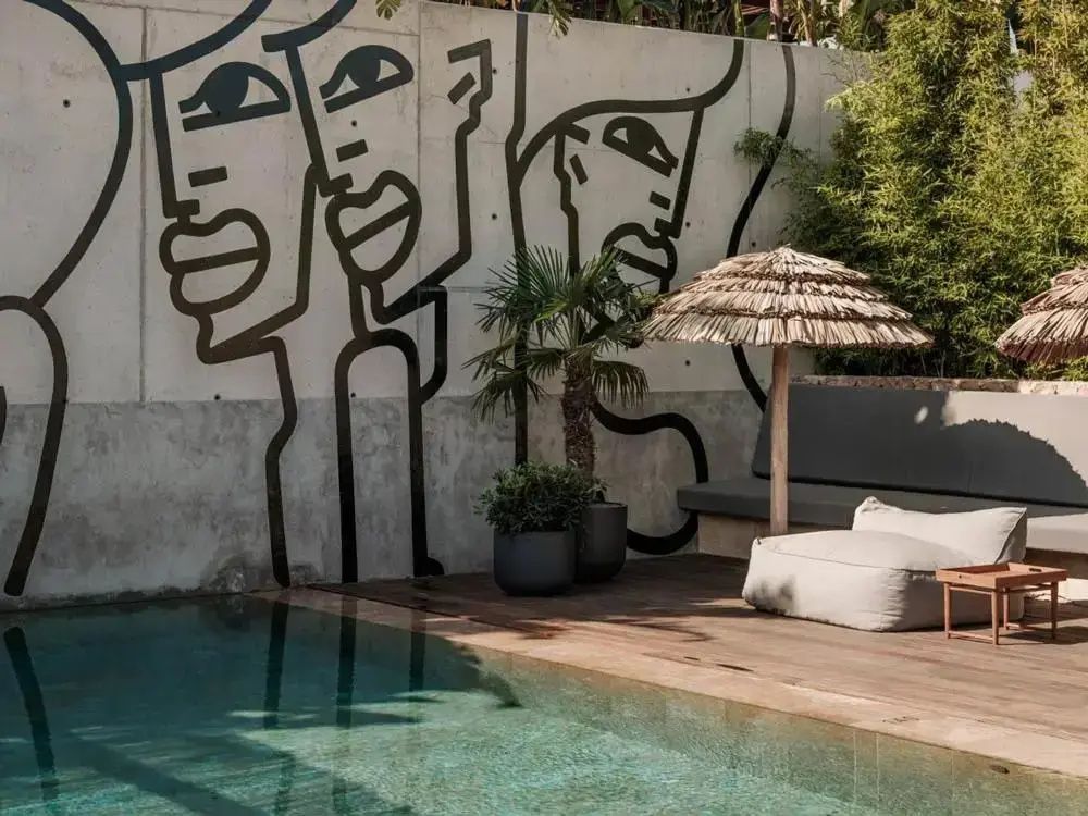 Poolkante mit Holzterrasse, Sitzkissen, Strohschirm und Wandzeichnung auf Betonwand.
