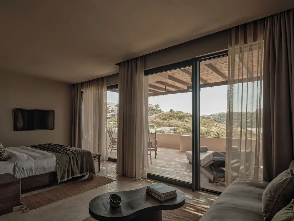 Ein helles Hotelzimmer mit Doppelbett, Couchtisch und Blick auf die sonnige Terrasse mit Hügellandschaft.