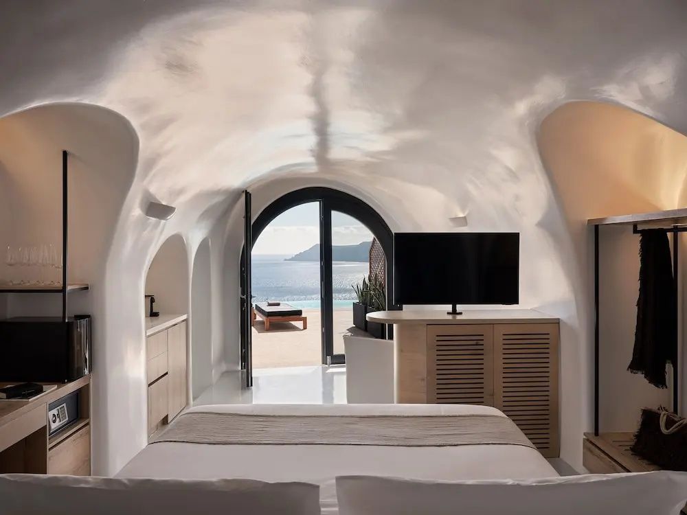 Weißes Höhlenzimmer mit Bett, TV und Blick auf das Meer durch die Terrasse.
