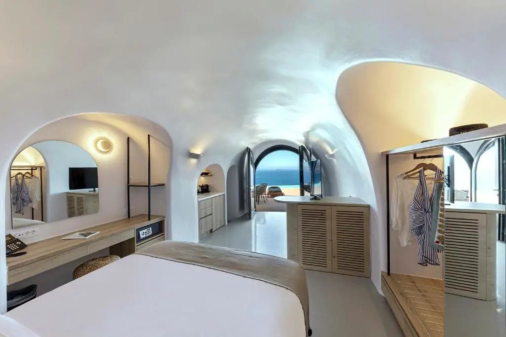 Weißes Höhlenzimmer mit Doppelbett und Blick durch die Bogentür auf das Meer.