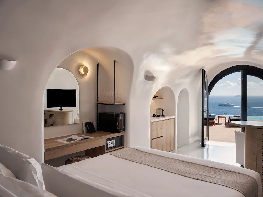 Höhlenzimmer mit Holztisch, Tresor, Minibar und Tür zur Terrasse mit Meerblick.