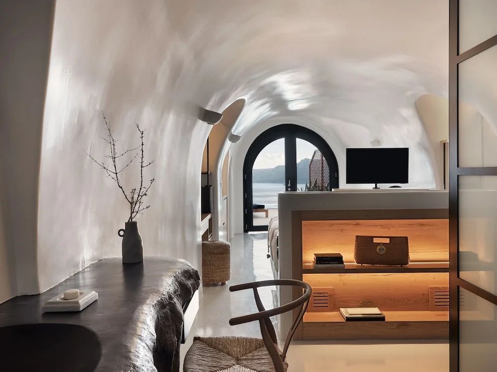 Weiß getünchte Höhlensuite mit Holzregal, Fernseher und Blick durch ein Bogenfenster aufs Meer.