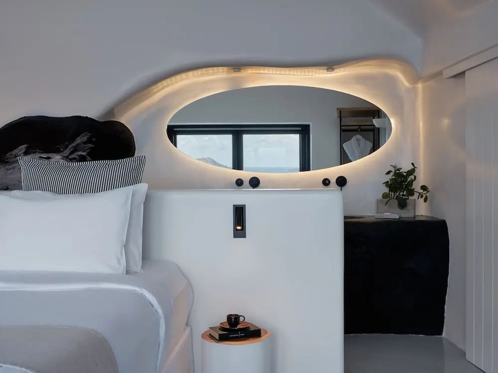 Schlafbereich mit weißem Bett, ovalem beleuchtetem Wandspiegel und modernem Nachttisch in Höhlenarchitektur.