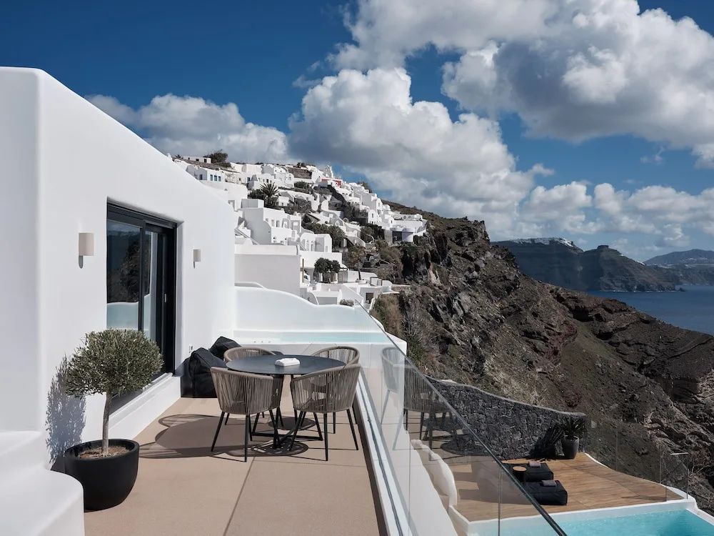 Terrasse mit Esstisch, Stühlen und Glasgeländer vor Felsküste mit traditionellen weißen Häusern.