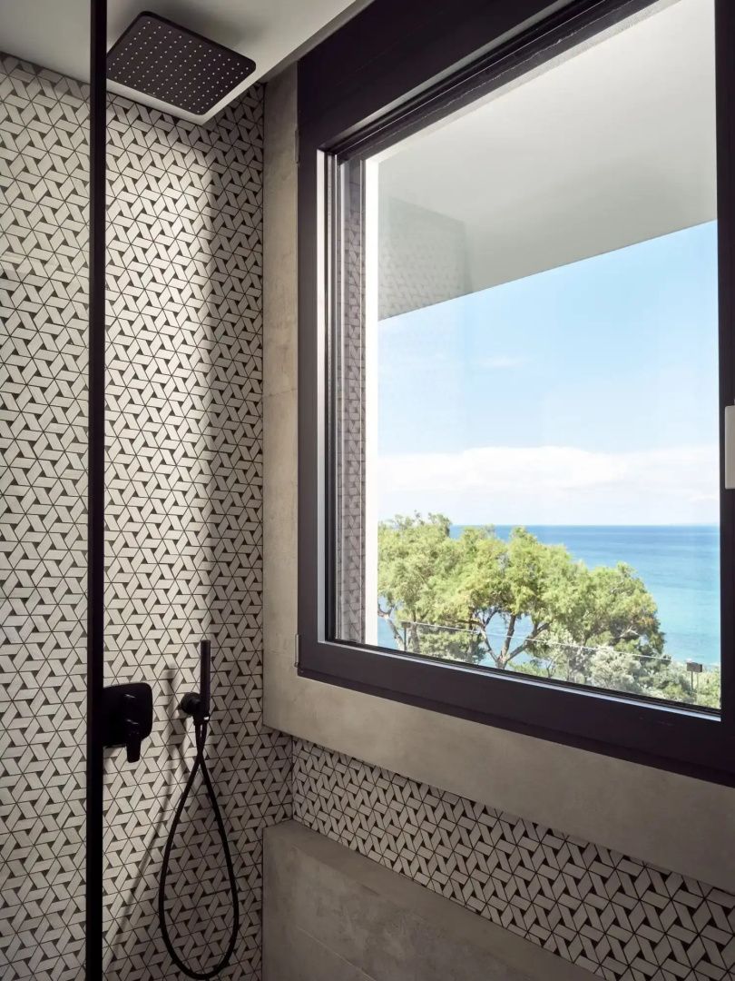 Blick durch ein Badezimmerfenster auf grüne Baumkronen und das blaue Meer.