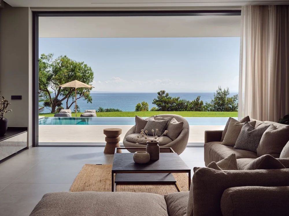 Wohnzimmer mit Sitzmöbeln und Blick durch die Glasfront auf Terrasse, Pool und Meer.