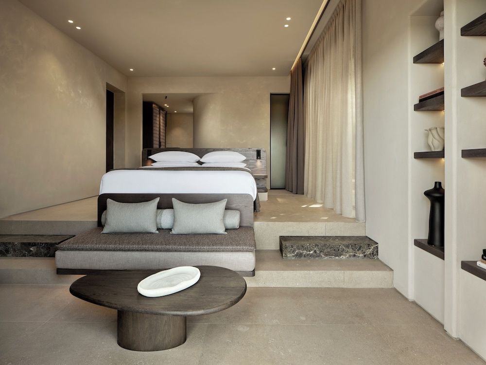 Helles Hotelzimmer mit organisch geformtem Couchtisch, Sitzbank und erhöhtem Bett.