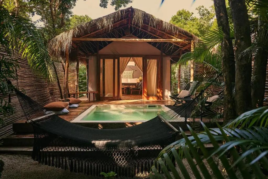 Bungalow mit Reetdach, kleinem Pool und einer schwarzen Hängematte im Freien.