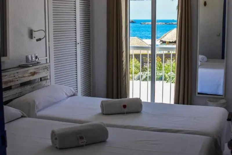 Helles Hotelzimmer mit zwei Einzelbetten und Blick durch das Fenster auf das blaue Meer.