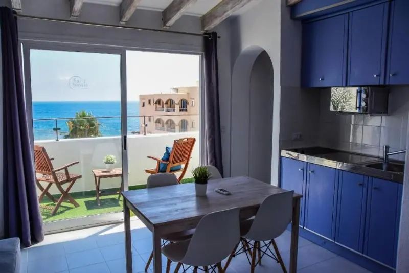 Wohnbereich mit Holztisch, blauer Küchenzeile und Balkon mit Aussicht auf das Meer.