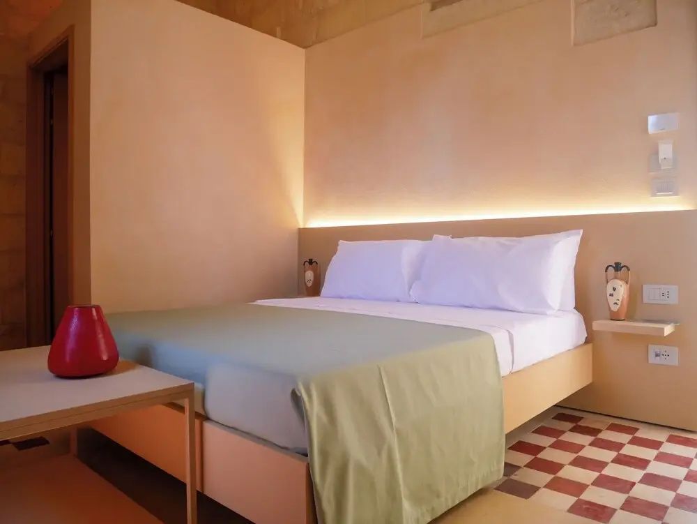 Doppelbett mit heller Bettwäsche, indirekter Beleuchtung und Holzrahmen in einem hellen Hotelzimmer.