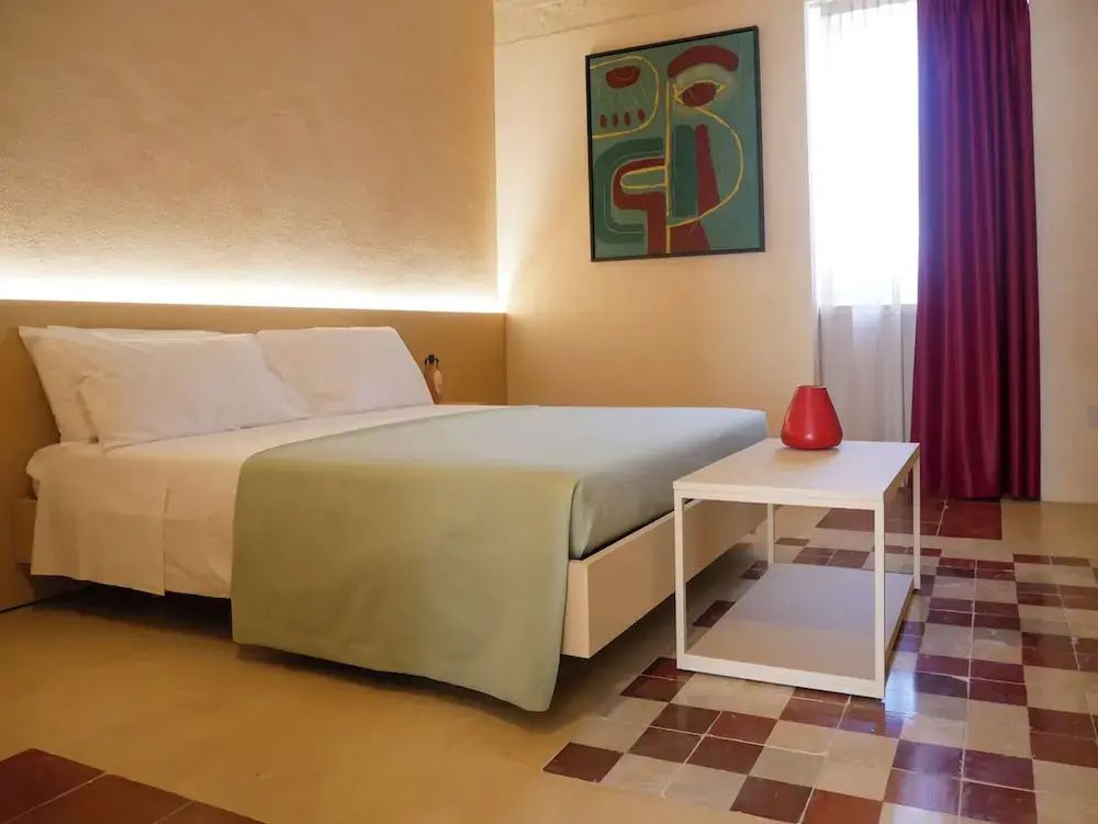 Schlafzimmer mit Doppelbett, abstraktem Wandbild, Fenster mit rotem Vorhang und Fliesenboden.