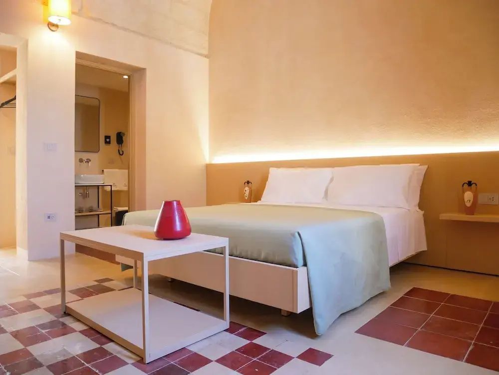 Hotelzimmer mit modernem Doppelbett, Tischchen und Blick in das angrenzende Badezimmer.