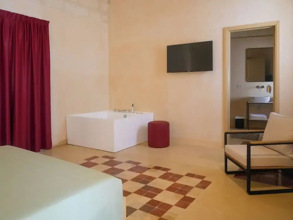 Hotelzimmer mit Whirlpool in der Ecke, rotem Hocker, Wandfernseher und gemütlichem Sessel.