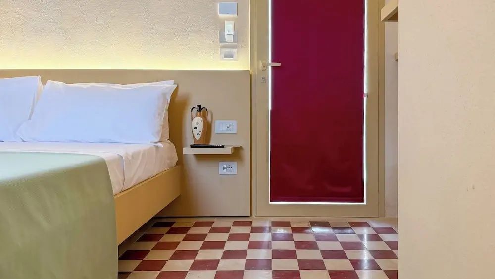 Blick auf das Bett, einen Nachttisch und rotweißen Fliesenboden.
