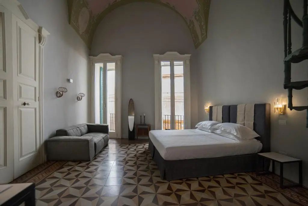 Großzügiges Hotelzimmer mit gewölbter Decke, Doppelbett, grauem Sofa und Fliesenboden.