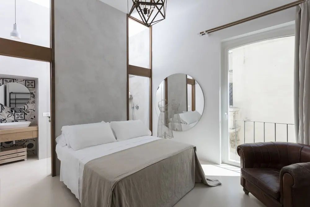 Zimmer mit weißem Doppelbett, einem hohen Fenster und einem großen runden Wandspiegel.