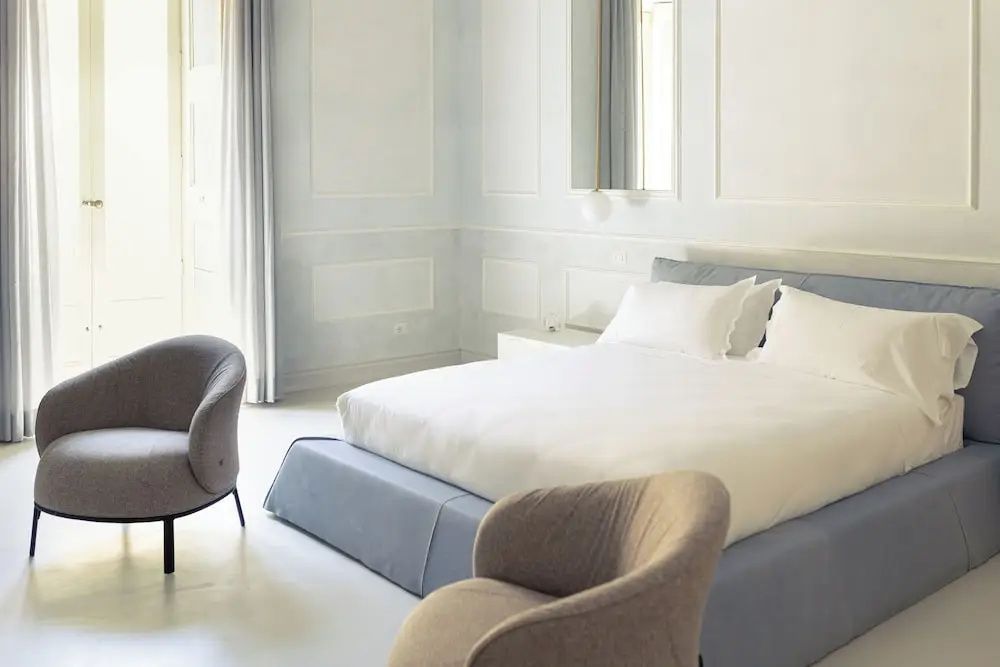 Hell eingerichtetes Hotelzimmer mit Doppelbett, weißer Bettwäsche und grauen Sesseln.