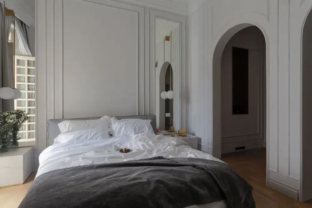 Doppelbett mit weißer Bettwäsche, grauer Tagesdecke und modernen Hängelampen im hellen Zimmer.