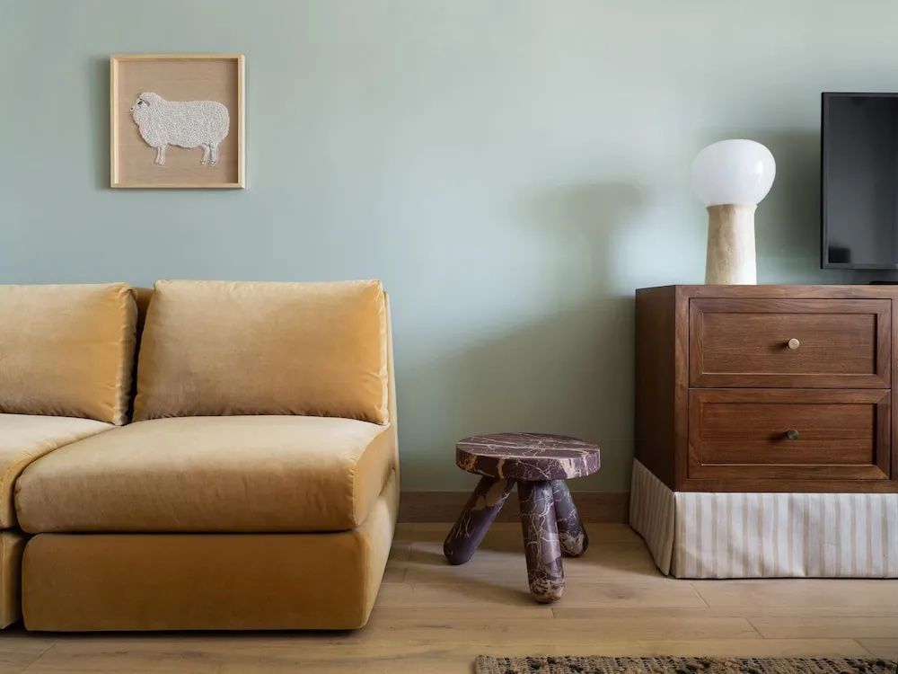 Gelbes Samtsofa neben einem kleinen Steinhocker und einer braunen Holzkommode.