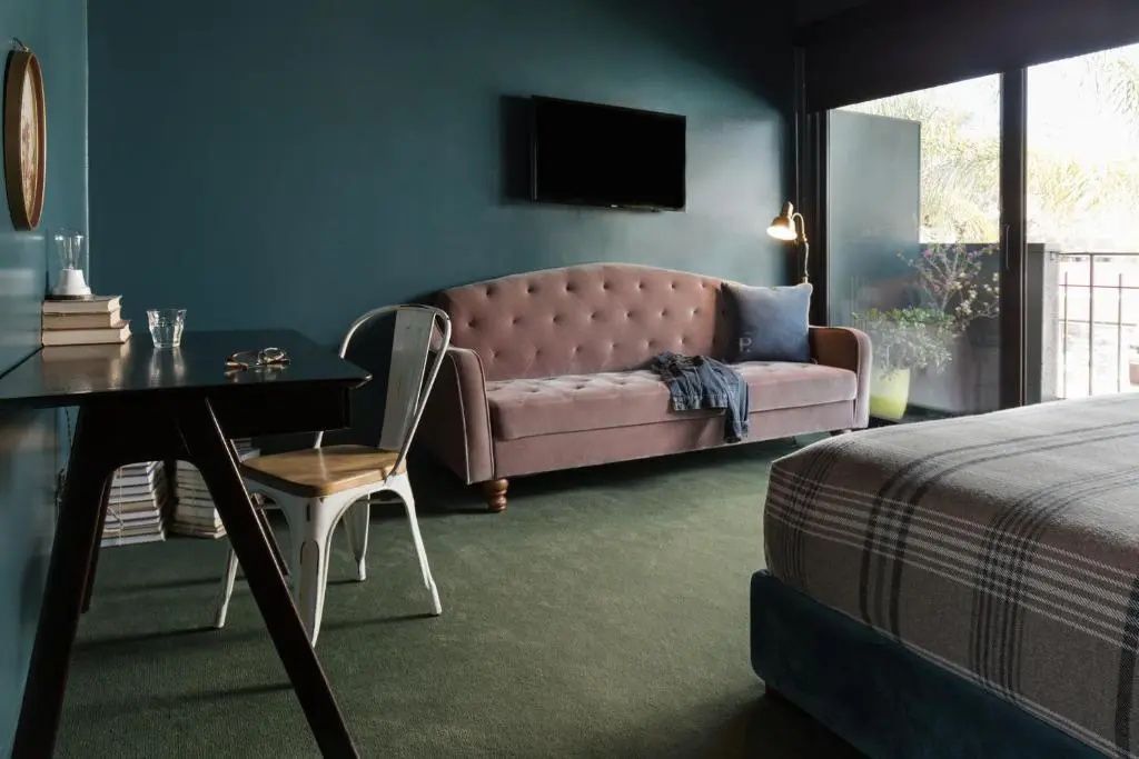 Sitzbereich im Hotelzimmer mit altrosa Samtsofa, Fernseher an der dunklen Wand und Schreibtisch.