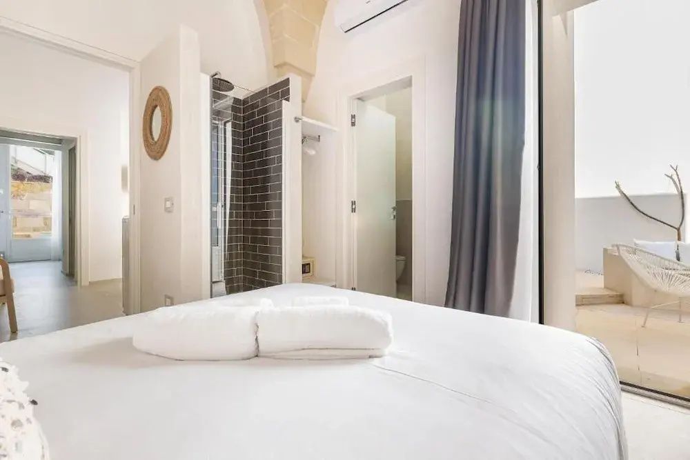 Schlafzimmer mit weißem Bett und Zugang zum Bad mit dunkler Dusche.