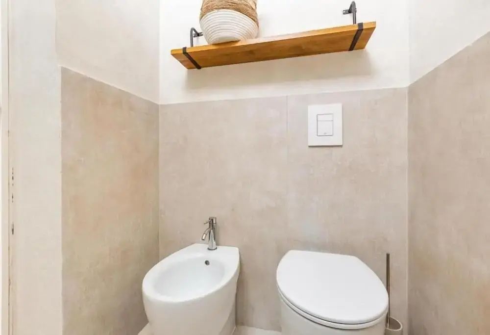 Weißes Bidet und Toilette vor beigen Fliesen mit kleinem Holzregal an der Wand.