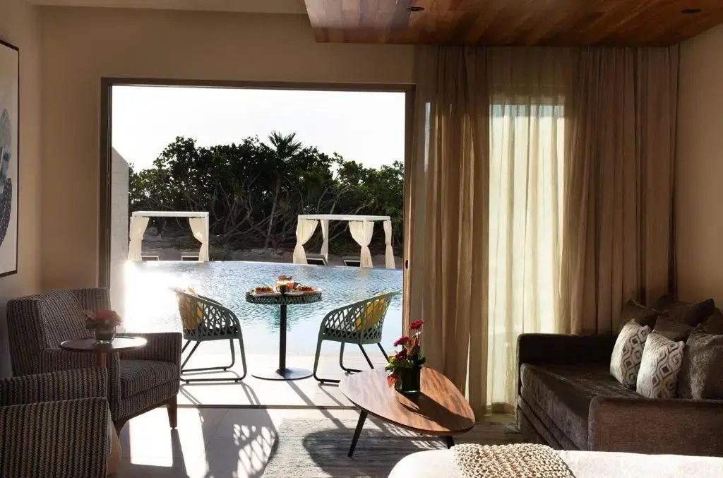 Blick aus dem Zimmer auf die Terrasse mit Sitzgelegenheiten und den dahinterliegenden Pool.