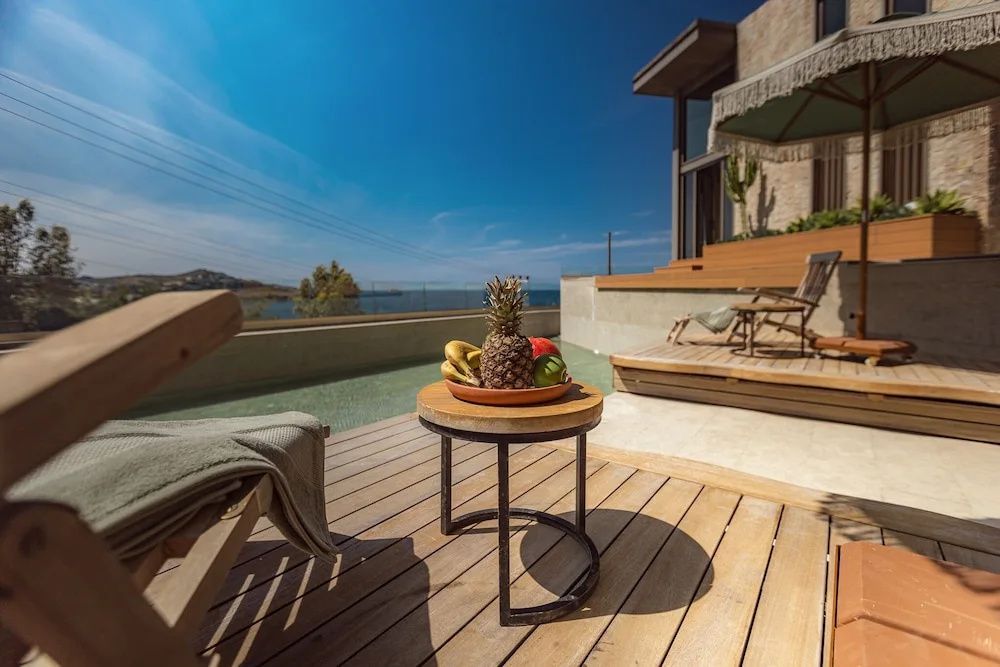Holzterrasse mit Liegestühlen und einem Tisch mit Obstschale vor dem Meer.