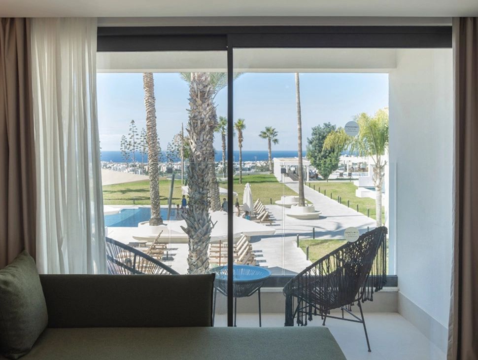 Blick durch eine Fensterscheibe auf die Hotelanlage mit Pool, Palmen und Meer im Hintergrund.