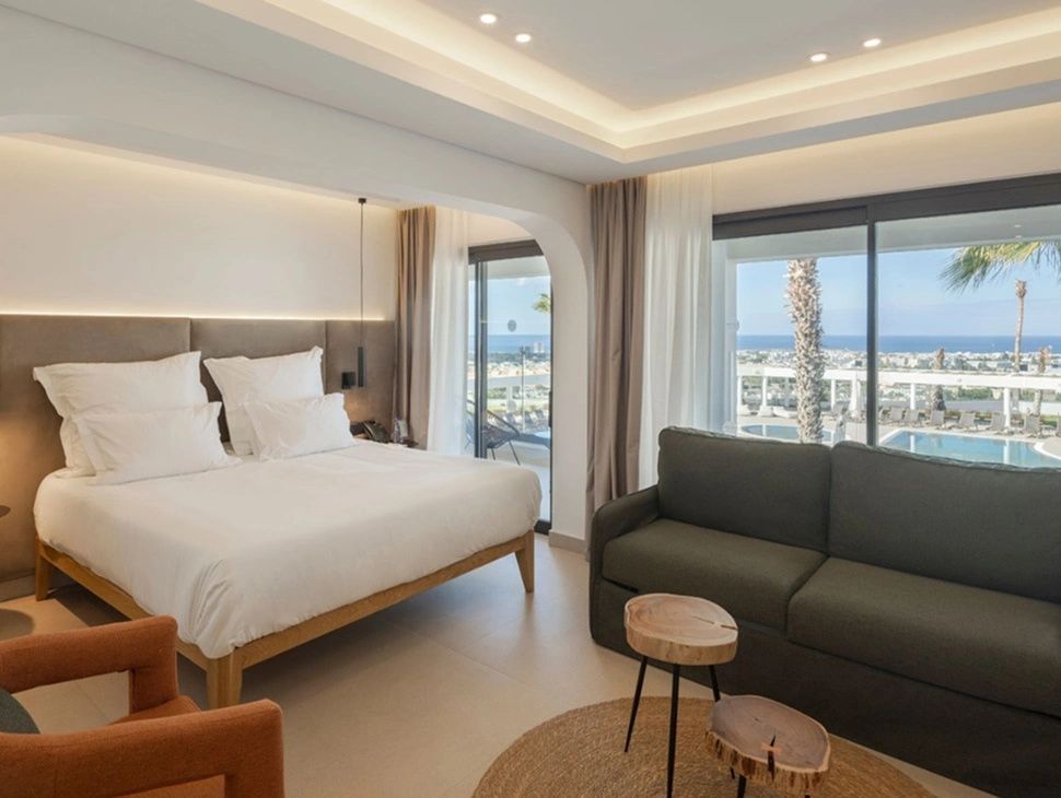 Helles Hotelzimmer mit Doppelbett, dunklem Sofa und Balkon mit Ausblick auf das Meer.