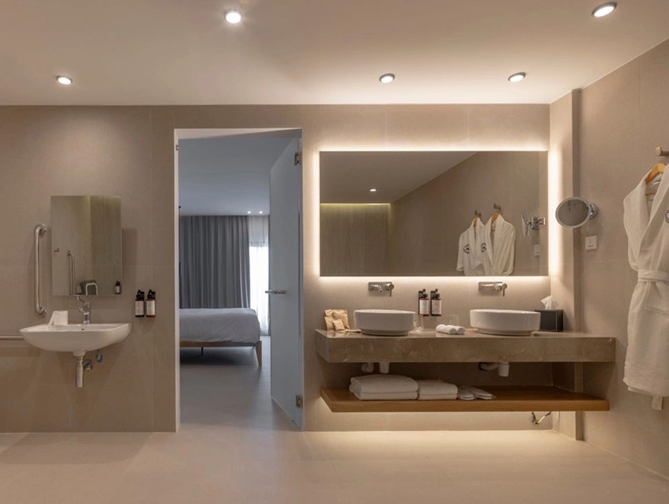 Badezimmer mit zwei Waschbecken, beleuchtetem Spiegel und Blick ins Schlafzimmer.