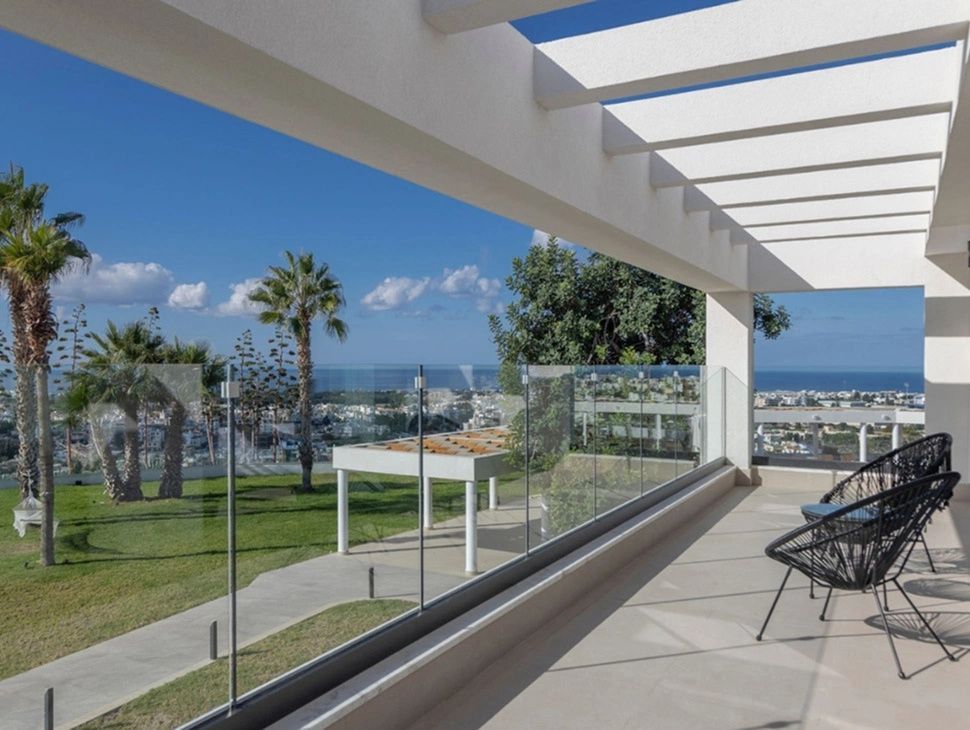 Terrasse mit Pergola und Glasgeländer mit Blick auf den Garten und das Meer.