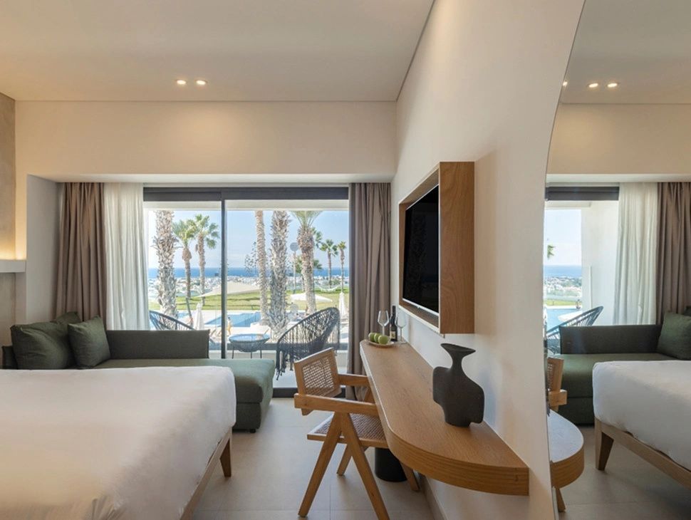 Helles Hotelzimmer mit Doppelbett, Schreibtisch und Glasfront zur Terrasse mit Meerblick.