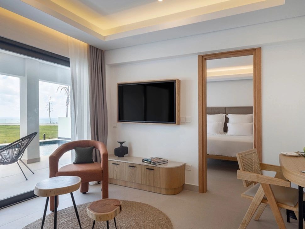 Modernes Wohnzimmer einer Hotel-Suite mit Fernseher, Sesseln und Durchgang zum Schlafzimmer.