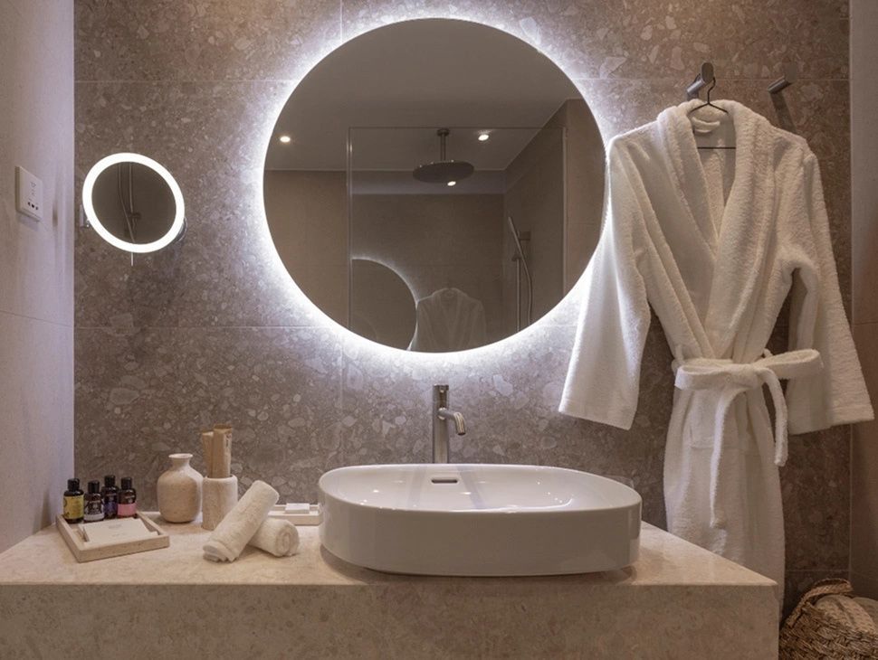 Modernes Badezimmer mit beleuchtetem Rundspiegel, Aufsatzwaschbecken und weißem Bademantel.