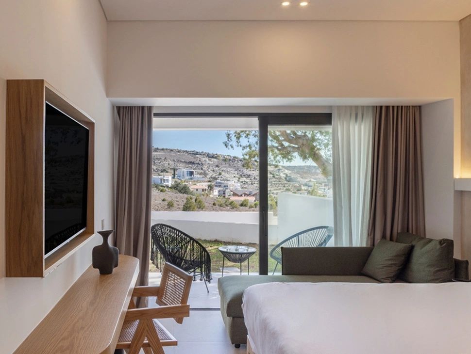 Helles Hotelzimmer mit Bett, Holzschreibtisch, Fernseher und Zugang zur eigenen Terrasse.