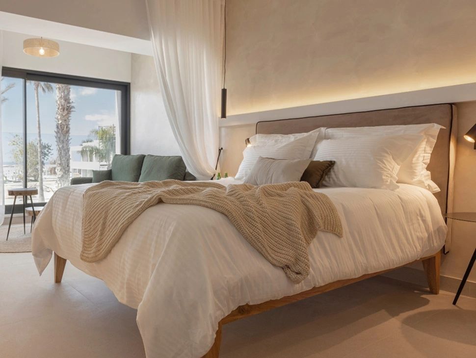 Hell eingerichtetes Hotelzimmer mit großem Doppelbett, Tagesdecke und Blick auf eine graue Couch.