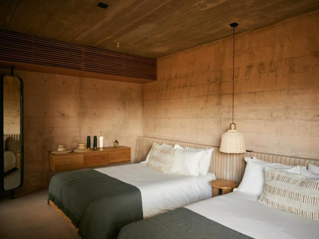 Schlafzimmer mit zwei Doppelbetten, Stampfbetonwänden und einer gewebten Hängelampe.