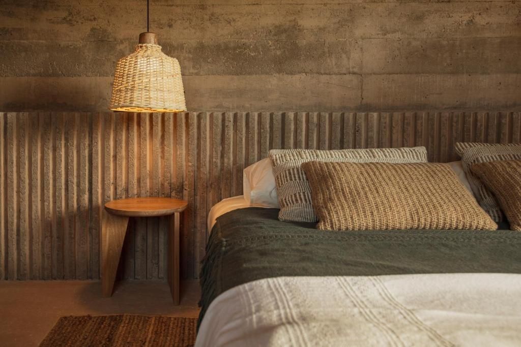 Bett mit Kissen und Tagesdecke neben einer geflochtenen Lampe und Betongestaltung.